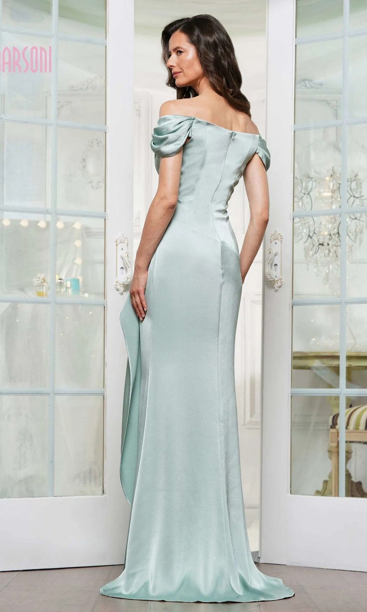 A woman stands indoors facing away from glass-paneled doors, wearing the Marsoni MV1351 Long Formal Dress—an off-the-shoulder, light blue evening gown with a fitted silhouette and slight train.
